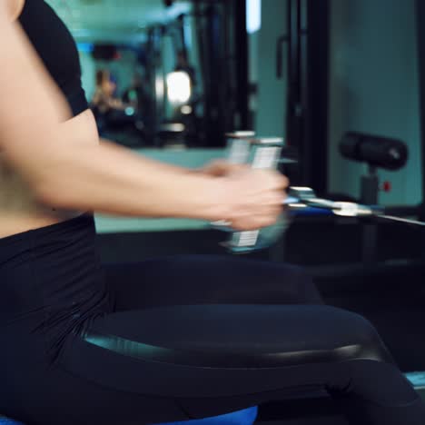 A woman is sitting behind a cardio trainer and working with her hands to strengthen the muscles of her arms and chest in the gym. Close-up. Blurred Background