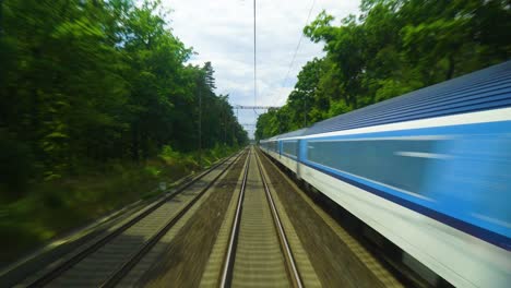 train on the railway tracks surrounded by lush green trees and a bright blue sky train is traveling opposite the track with power lines above it tall trees are casting shadows onto the ground below