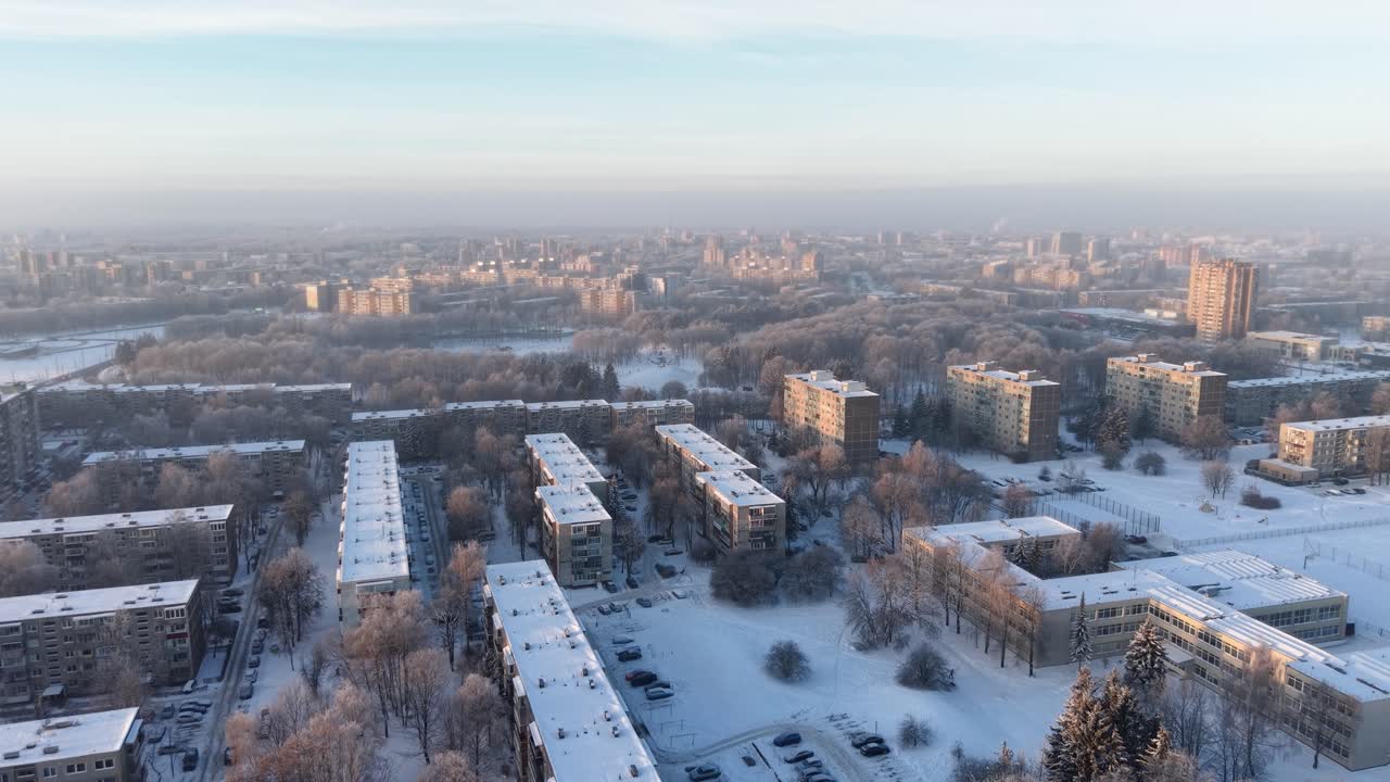 Drohnenvideo, das eine friedliche, schneebedeckte städtische Nachbarschaft im Morgengrauen einfängt. Luftaufnahmen, die geordnete Stadtblöcke, frostige Bäume und eine dunstige Skyline unter sanftem Morgenlicht zeigen