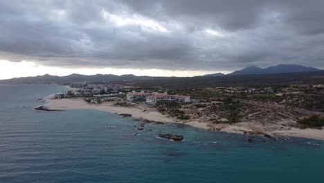 Vista-Aérea-Sobre-La-Hermosa-Costa-De-La-Playa-El-Chileno-Baja-California-Sur,-México-Con-Vista-Al-Mar-Azul,-Edificios-Hoteleros-Y-Paisaje-Con-Exuberante-Vegetación-Con-Magníficas-Cadenas-Montañosas