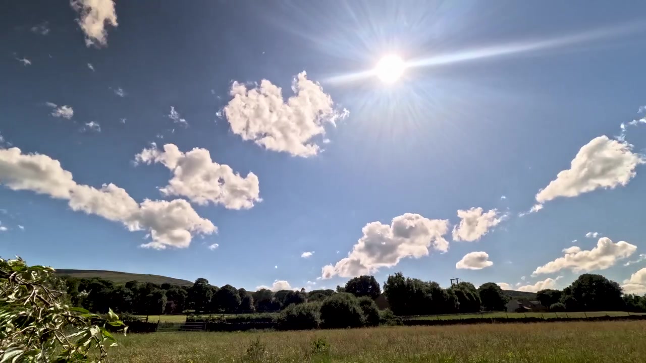 Fast-moving white clouds stream past the bright sun above a green English countryside field in a summer sky timelapse.