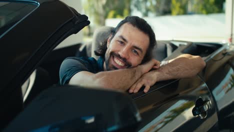 A-young-brunette-guy-looks-out-of-the-open-window-of-a-dark-gray-convertible-and-clouds-on-it,-smiles-and-looks-at-the-camera