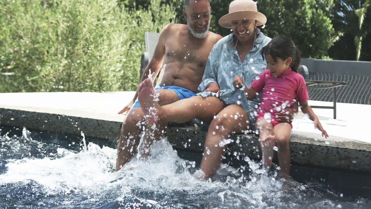 abuelos biraciales con su nieta disfrutan de salpicaduras de agua junto a una piscina