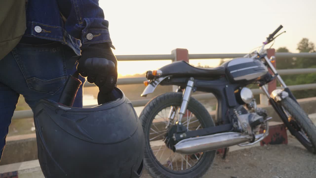 Motorcyclist holding helmet on a bridge at sunset