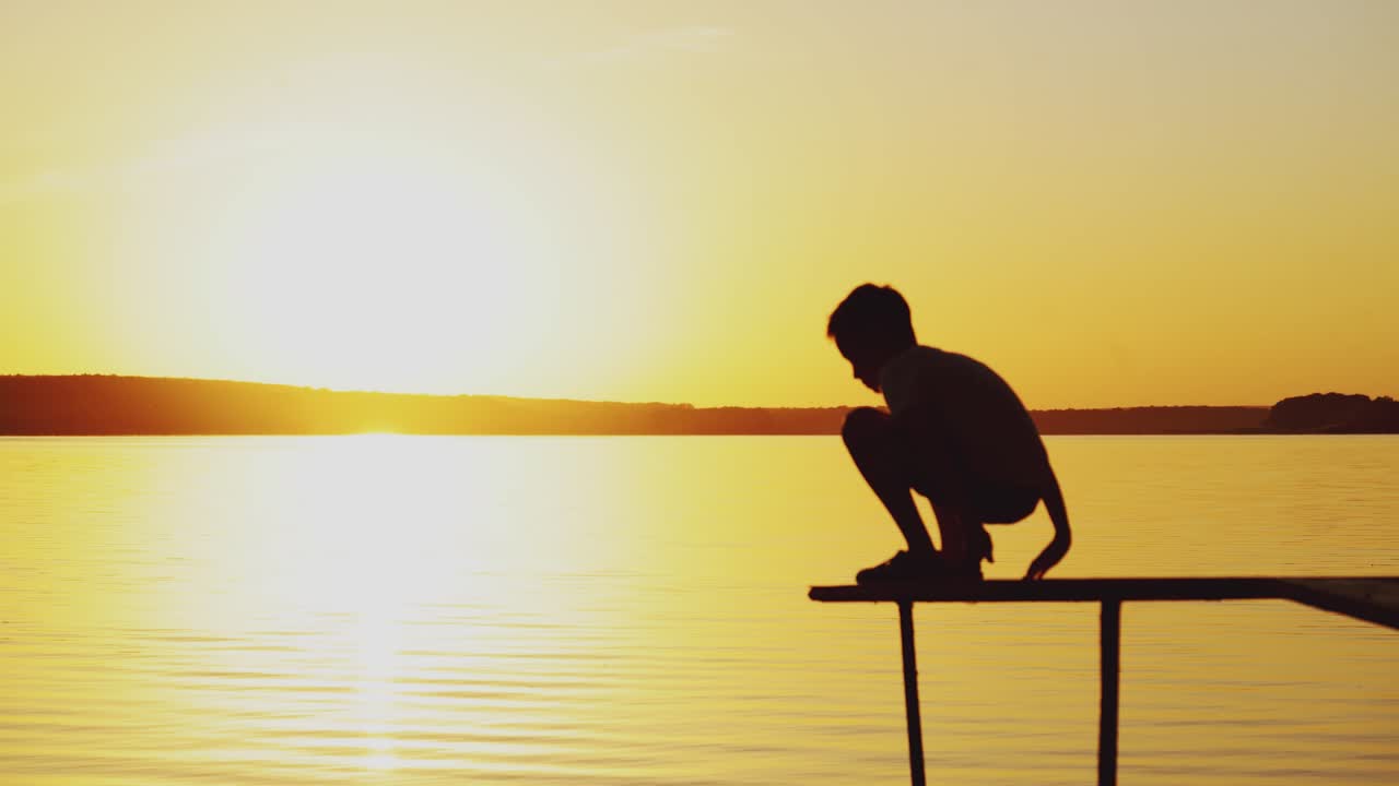 a little boy is sitting in a lotus position on the edge of a bridge above a river at sunset in the summer.