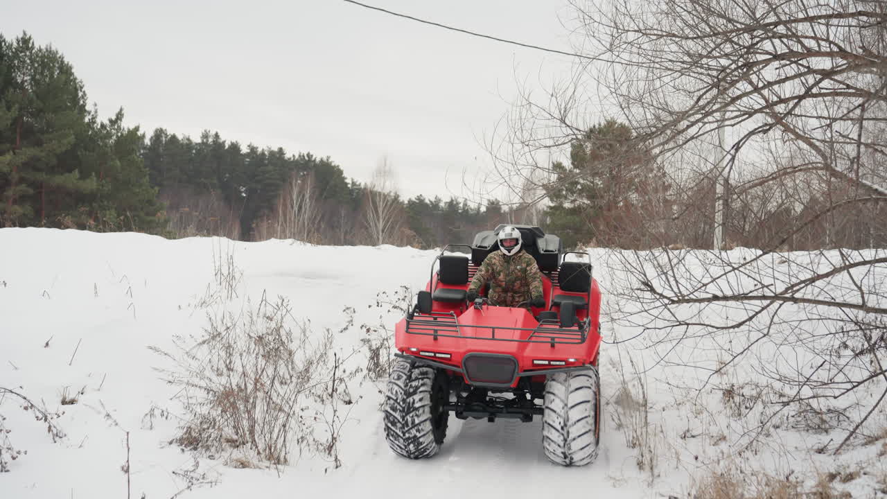 ATV rosso che sfreccia lungo un sentiero innevato, primo piano frontale con pneumatici sovradimensionati che sollevano neve. Il pilota, con giacca invernale e casco, naviga attraverso un campo aperto verso la telecamera, con una pineta.