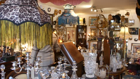 a person in a plaid coat browsing an antique shop filled with vintage glassware, ornate lampshades, and decorative items. the eclectic mix of collectibles and framed art creates a nostalgic atmosphere
