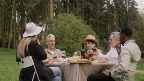 grupo de amigos de mediana edad comiendo y hablando entre ellos sentados en la mesa durante una fiesta al aire libre en el parque 2