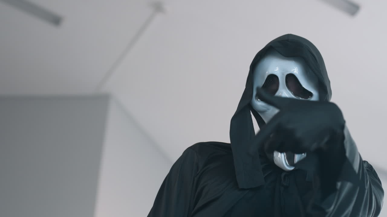 masked figure in black cloak raises gloved finger to lips in eerie silent gesture against pale studio backdrop with mirror reflection hinting hidden presence