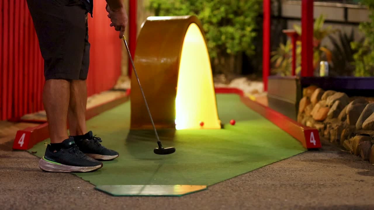 A person attempts a putt on a mini golf course at night, illuminated by warm lighting, creating a playful atmosphere