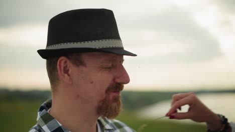 a close-up shot of a man wearing a hat, gently smiling as a woman's hand playfully touches his nose with a blade of grass. the background is blurred