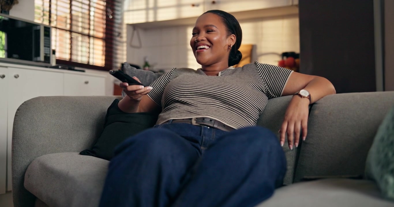 Glad ung kvinde, der slapper af på sofaen og ser fjernsyn derhjemme
