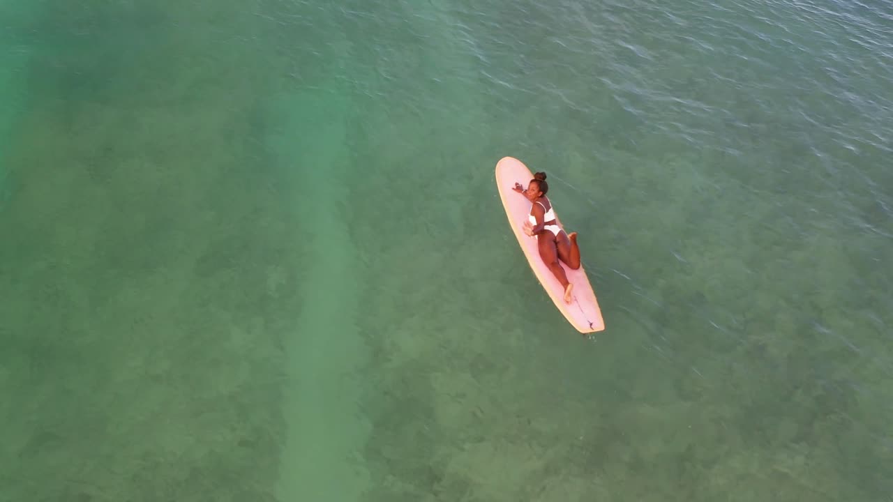 Female surfer paddles across shallow reef waters in Hawaii captured from a top-down drone view. The crystal-clear water reflects sunlight, showcasing serene island vibes and nature's beauty