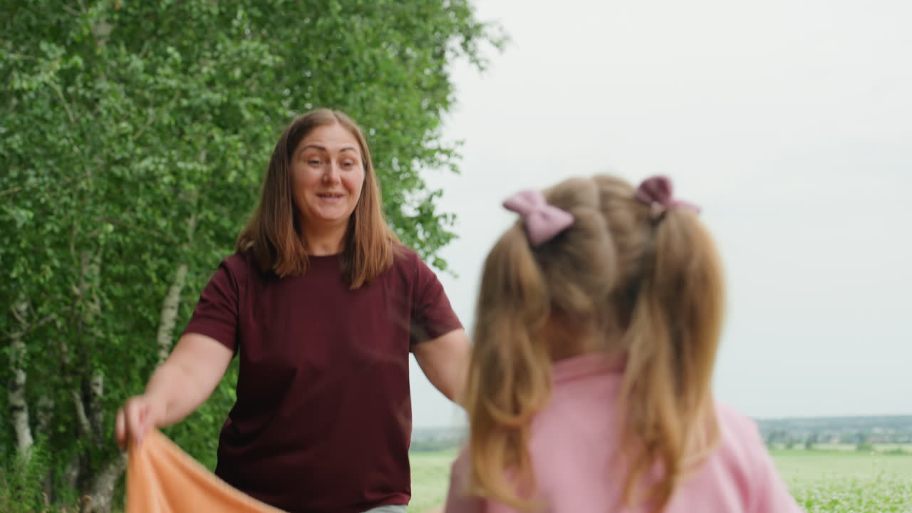 하얀 블랭킷을 던지며 활짝 웃고 머리 위로 천을 잡는 백인 엄마와 딸, 장난기 넘치는 피크닉 분위기, 엄마의 미소, 나뭇잎이 우거진 나무 배경, 따뜻한 햇살과 가까운 가족의 유대감