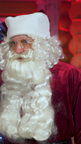 portrait of a real santa with white beard. senior man in santa clause costume wearing glasses standing in christmas decorated room and talking. vertical video