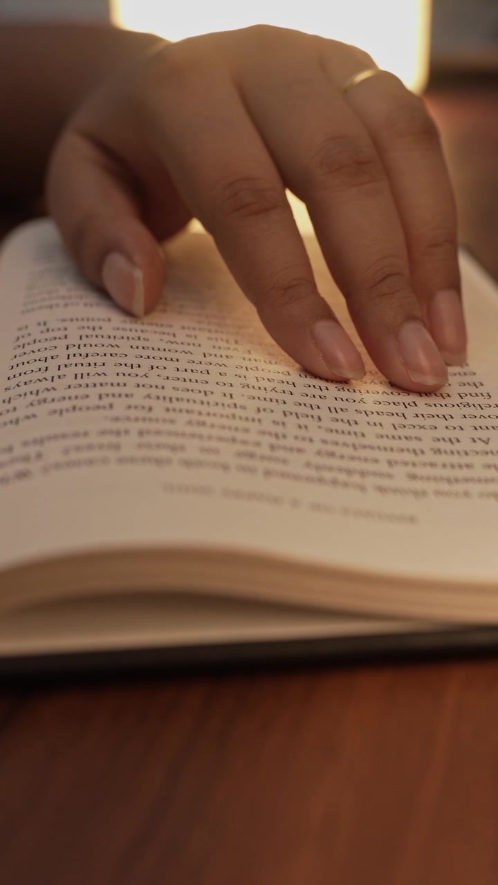 Primo piano della mano di una donna che preme e leviga delicatamente una pagina di un libro aperto mentre legge, con testo stampato chiaramente visibile, superficie di un tavolo di legno caldo e luce calda che illumina