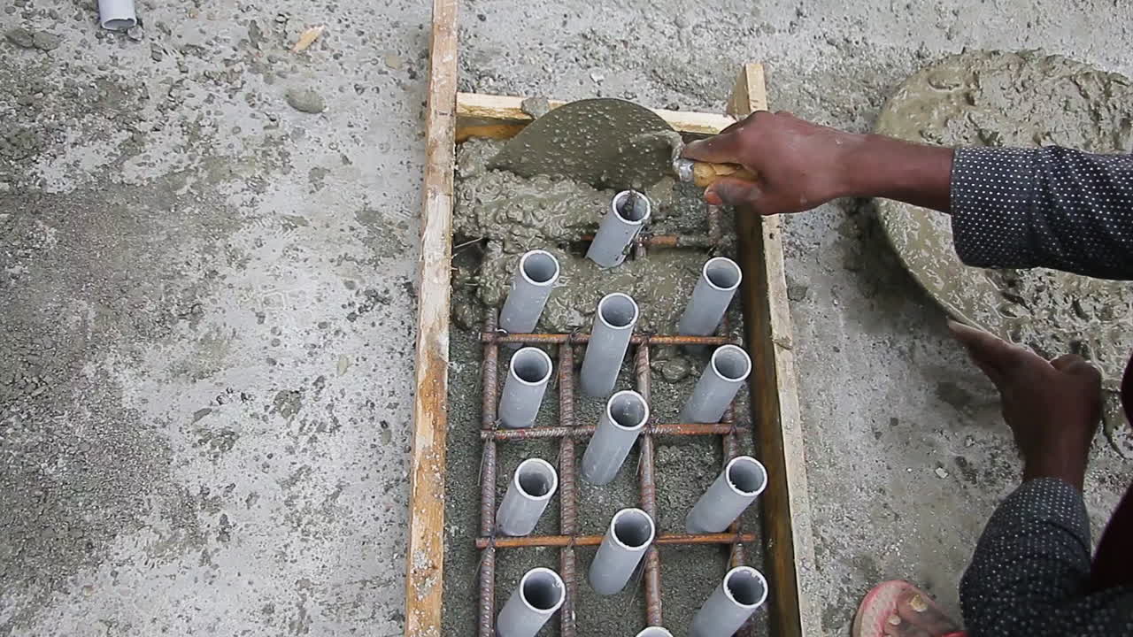 Indian mason worker placing concrete mix into slab mold and leveling it around PVC pipes for construction