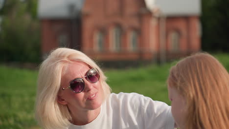 young mother with sunglasses chatting with daughter outdoors during sunset, warm light on faces, soft blur background, affectionate connection and relaxed conversation in green park setting