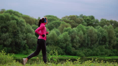 a girl in a pink jacket and black pants runs near the river in headphones preparing for the marathon