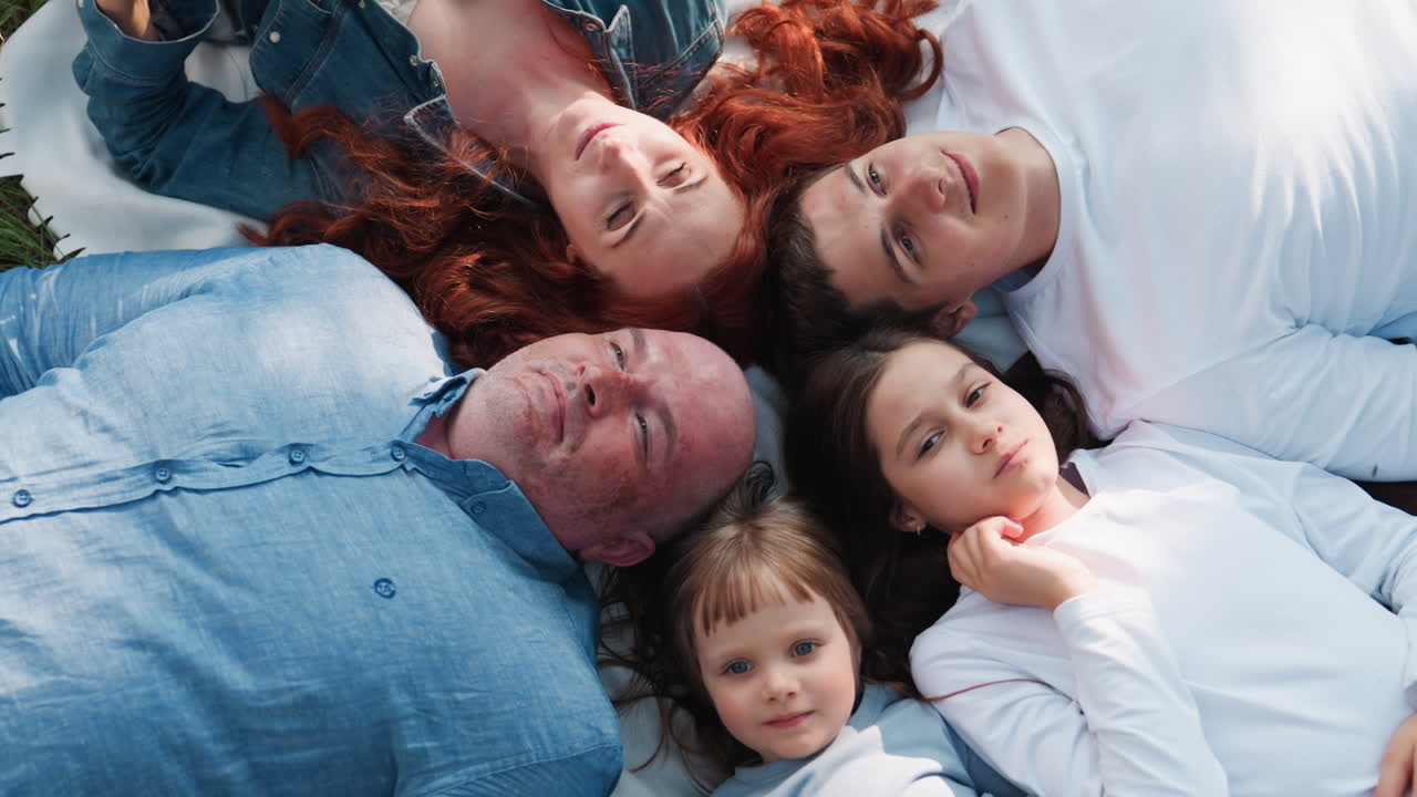 Aerial view of parents and kids lying faced up on picnic blanket outdoors, enjoying calm relaxation and family unity under sunlight, expressing peace, and tenderness in warm natural atmosphere