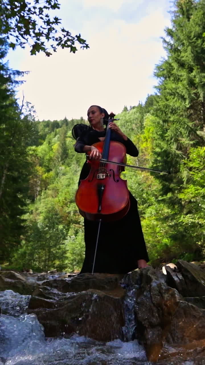 Luxury woman with musical instrument outdoors. Female musician performing music with a cello on amazing nature background with fresh mountain water. Vertical video
