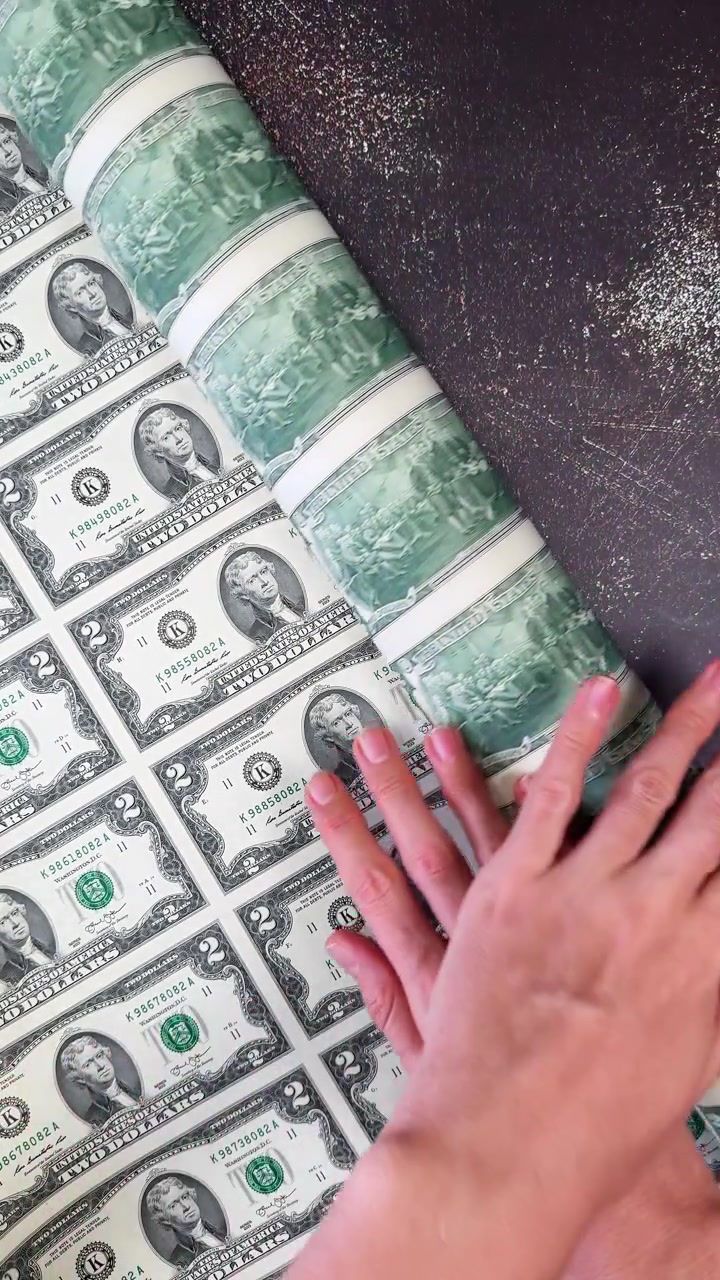 Rozwijanie arkusza pieniędzy. Widok z góry na kobiece dłonie rozwijające duży, niepocięty arkusz prawdziwych dwudolarowych banknotów amerykańskich na ciemnej, teksturowanej powierzchni.