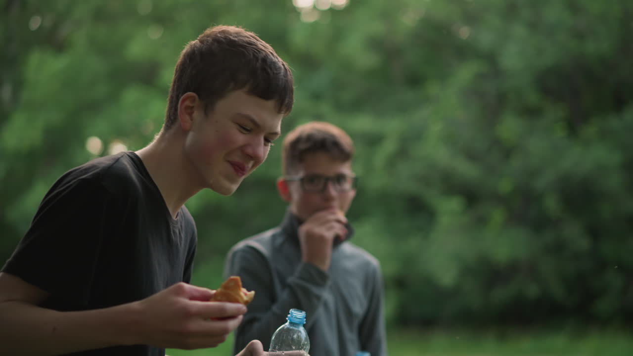 молодой мальчик в черном топ вспыхивает в смех, пьючи воду и держа закуску, с его другом на заднем плане чистит рот и смотрит, как он смеется, окруженный зеленью