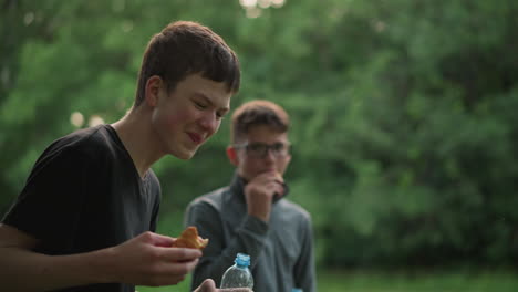 young boy in black top bursts into laughter while drinking water and holding a snack, with his friend in the background cleaning his mouth and watching him laugh, surrounded by a greenery