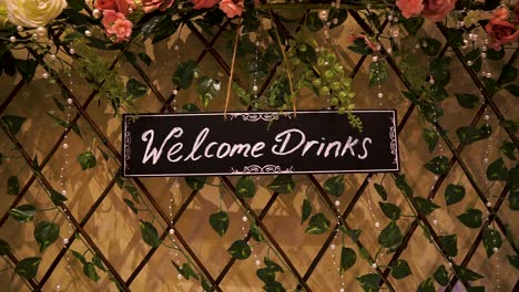 welcome drinks black background sign hanging in a fence that is covered with pink and yellow roses leaf's and pearls on asian wedding cinematic beautiful scenery