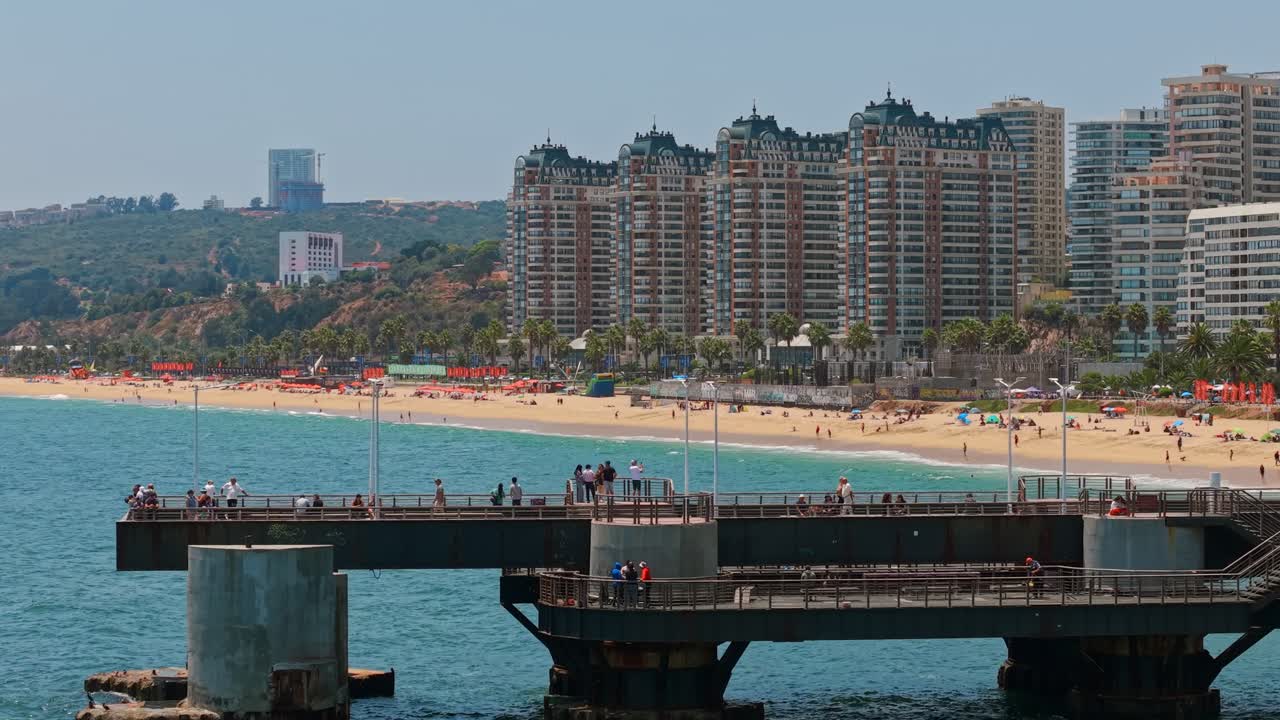 Bird's-eye view establishing telephoto dolly out of Vergara Pier in Viña del Mar on a sunny day, exclusive Puerto Pacifico condominiums on the beachfront