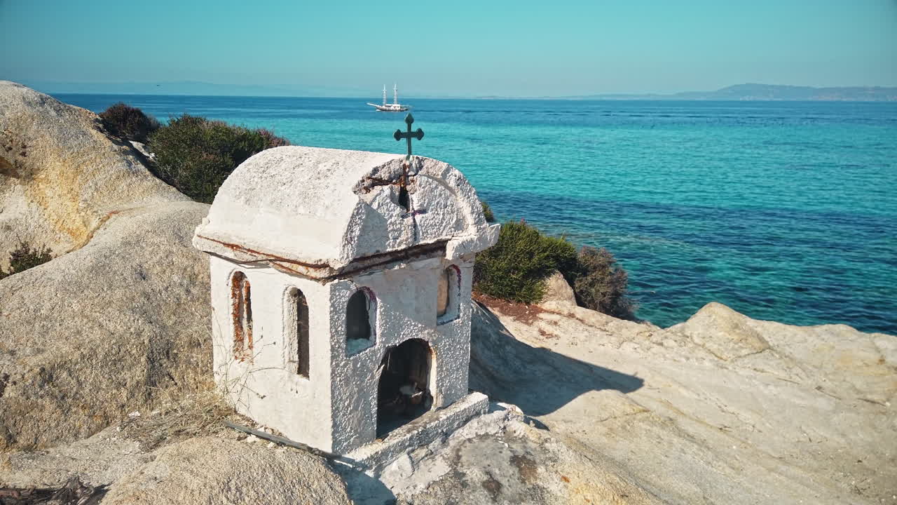 A small shrine located on rocks near the Aegean sea coast, water and moving yacht on the background, Greece. Slow motion
