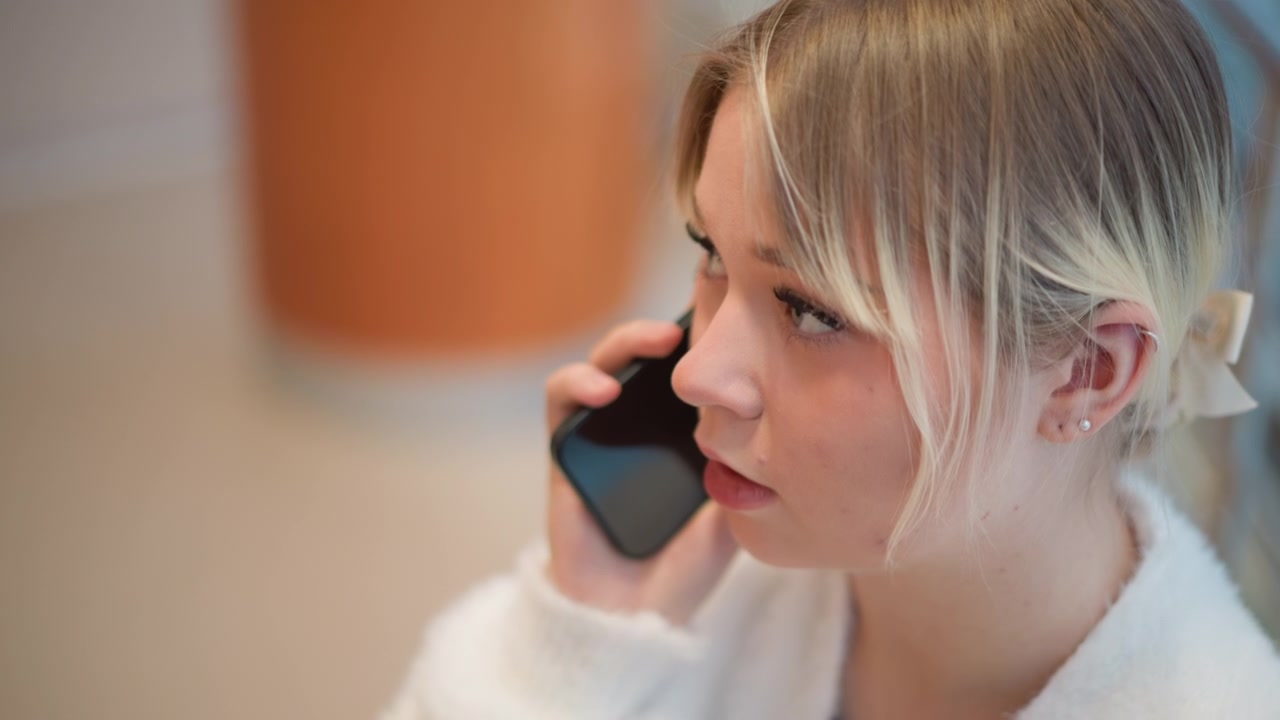 Zbliżenie młodej kobiety w szlafroku, trzymającej smartfon przy uchu, lekko pochylonej i przechylającej głowę ze skupionym wyrazem twarzy, zaangażowanej w spokojną rozmowę telefoniczną w pomieszczeniu.