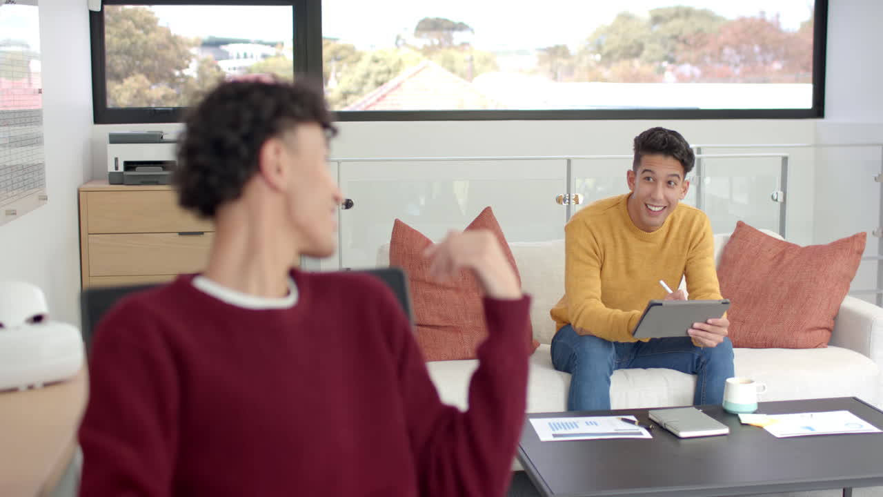Smiling man holding tablet, talking to partner sitting on couch at home