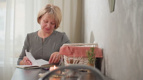 Lady focused on study flipping notebook backward thoughtfully with phone on desk near candle light cozy interior with natural daylight flowing through transparent curtains and basket
