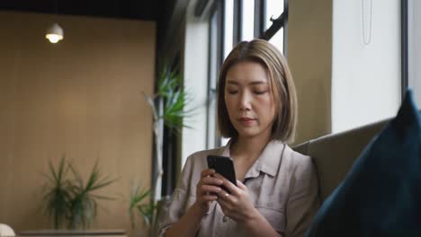 Mujer-De-Negocios-Asiática-Sentada-Junto-A-Una-Ventana-Usando-Un-Teléfono-Inteligente-En-Una-Oficina-Moderna