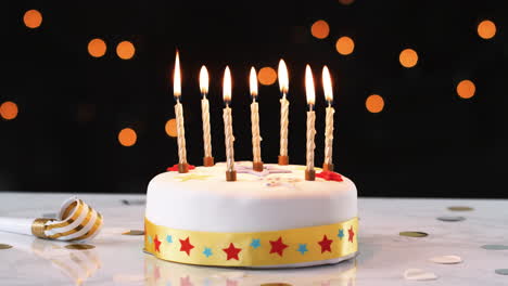 siete velas encendidas en un pastel de cumpleaños blanco y decorado, un soplador de fiesta a su lado, luces bokeh en el fondo