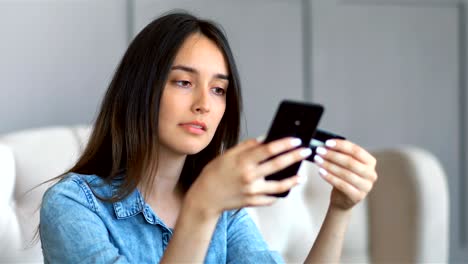 young pretty woman buying online with credit card and smartphone sitting on a couch in the living room at home