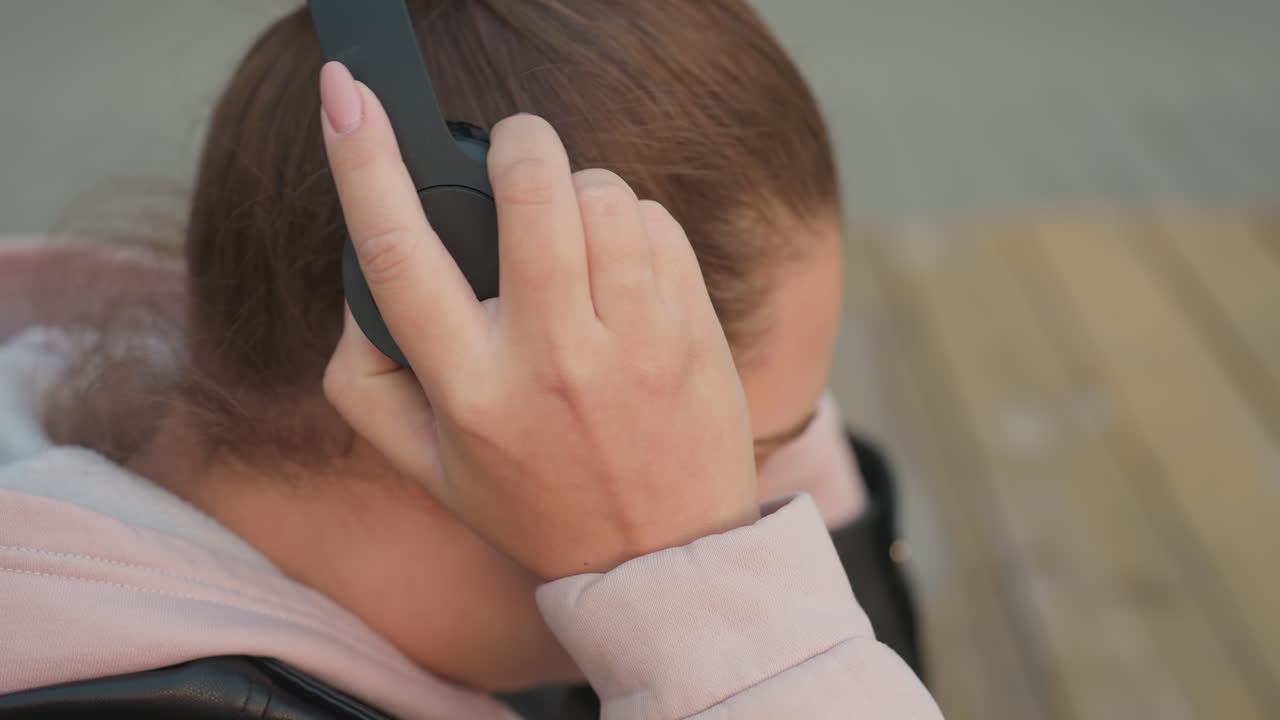 Close up young woman with polished nails wearing pink hoodie and black leather jacket seated at wooden table slowly lifts headset while gentle breeze moves her hair