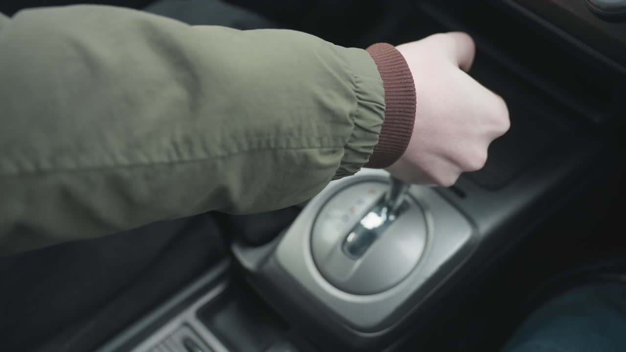 Close up top down view of driver pushing gear shift lever into park position in car center console showing hand grip on leather knob and metallic base under soft daylight in modern interior