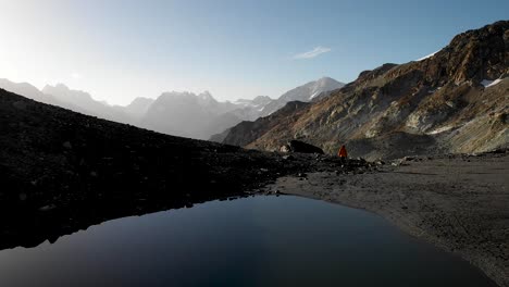 Aerial-flyover-over-an-alpine-lake-near-Arolla-in-Valais,-Switzerland-with-a-perfect-reflection-and-a-hiker-walking-alongside-on-a-sunny-early-morning-in-the-Swiss-Alps