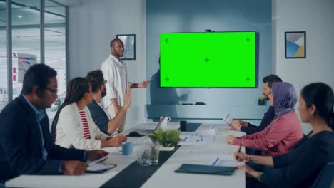 office conference room meeting presentation: black businessman talks, uses green screen chroma key wall tv. successfully presenting a product to group of multi-ethnic investors. e-commerce strategy