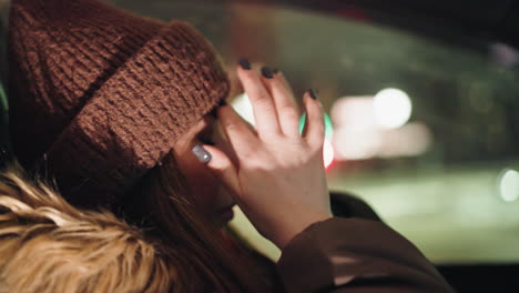 sicht aus der ersten person einer frau in einem wintermantel mit einer pelzkapuze, nachts in einem auto. sie passt ihren kopfwärmer mit schwarz bemalten fingern auf ihren kopf an. der hintergrund zeigt verschwommene stadtlichter