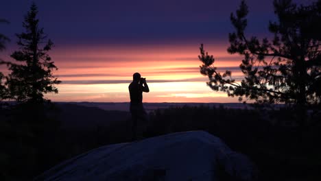 silhouette of male hiker photographer standing on a nordic mountain cliff looking towards horizon with a summer sunset or a sunrise over a big lake view
