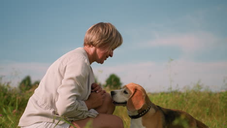 pet lover squatting outdoors with her dog in vast grassy field on sunny day, interacting playfully as the dog looks up with tongue out happily jumps up and kissed her, surrounded by lush nature