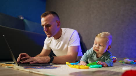 caucasian dad and infant son lie on the floor. father works on laptop. baby boy looks at his toys.