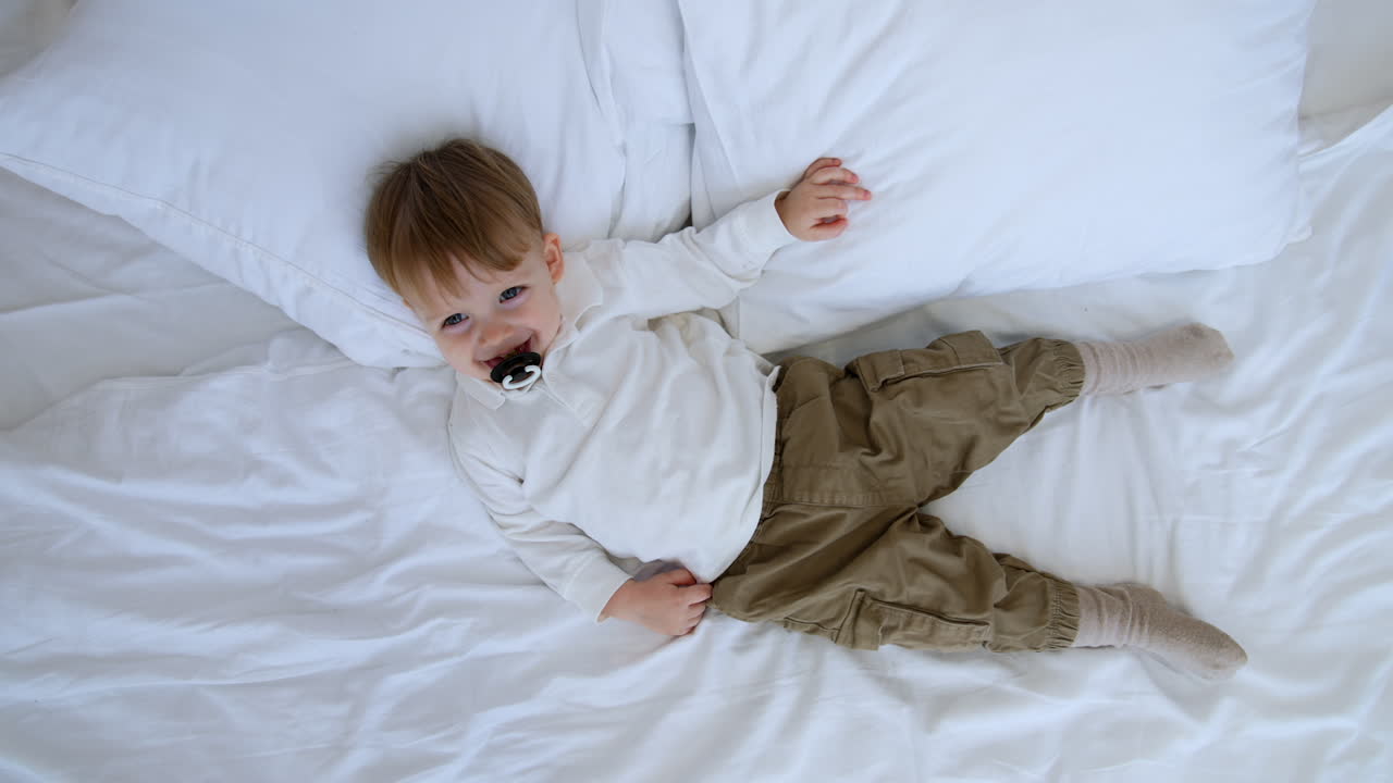 Lovely toddler boy in white shirt and grey pants lies on the big bed. Cute kid with pacifier smiles to camera. Top view.