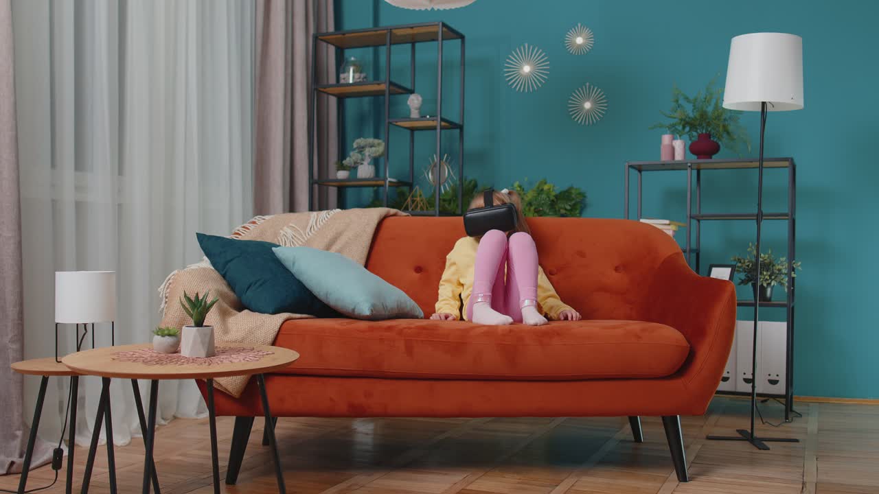 가정 소파에 앉아있는 유아 소녀가 시레이션 게임을 하기 위해 가상 현실 헤드 헬 앱을 사용합니다.