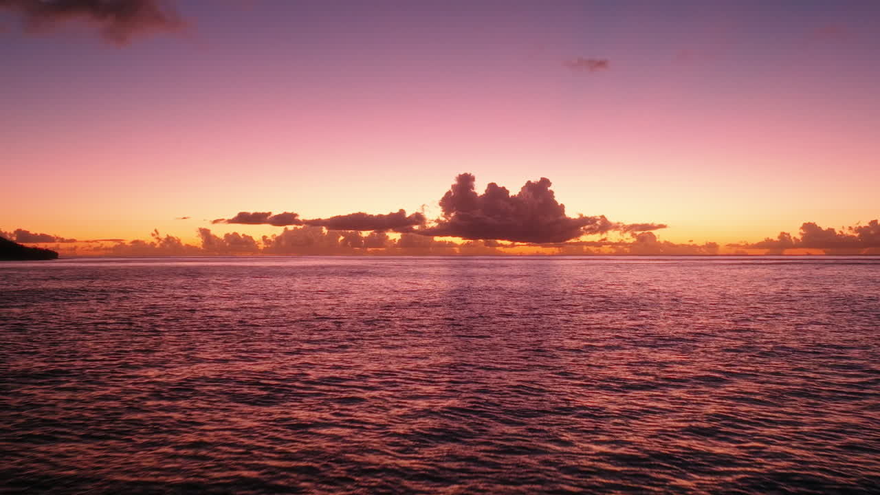Drone flying forward over the ocean at sunset, passing a dark coastal silhouette under an orange and purple sky