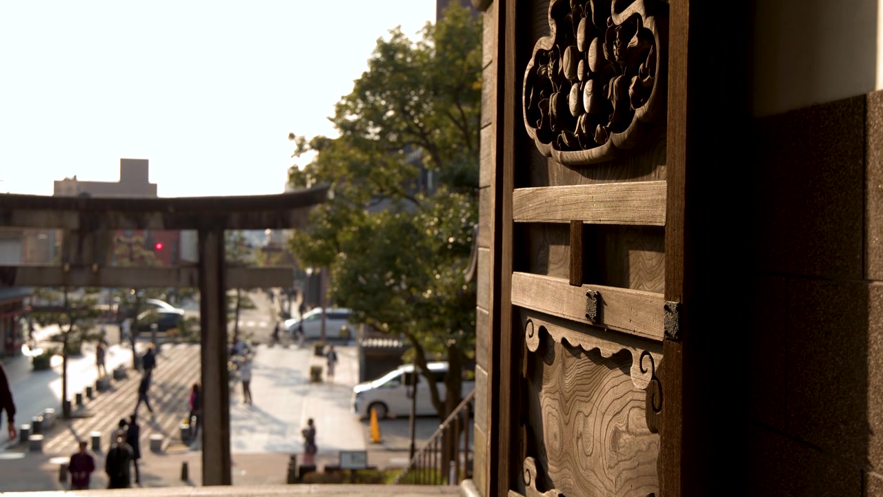 Intricate wooden carvings frame a view of a stone torii gate and busy city street