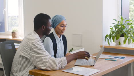 Junger-Arbeiter,-Der-Mit-Einem-Laptop-Arbeitet,-Der-An-Seinem-Schreibtisch-Sitzt,-Während-Eine-Muslimische-Geschäftsfrau-Mit-Ihm-Spricht,-Während-Er-Sich-Notizen-Macht-1